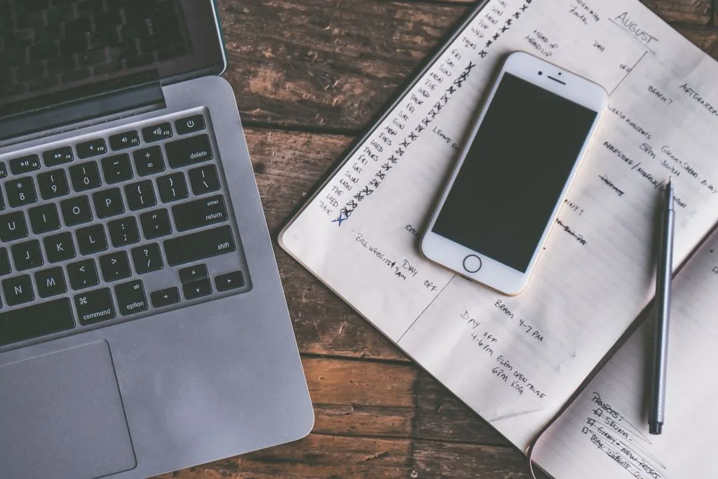 An open notebook beside a laptop and phone on a wooden desk, used for planning freelance work and admin