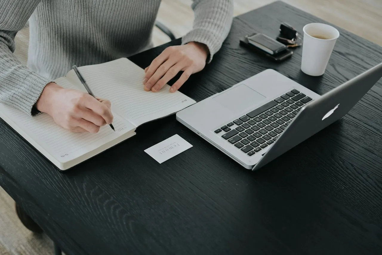 Student writing in a notebook at a desk with a laptop and coffee while learning how to write a scholarship essay