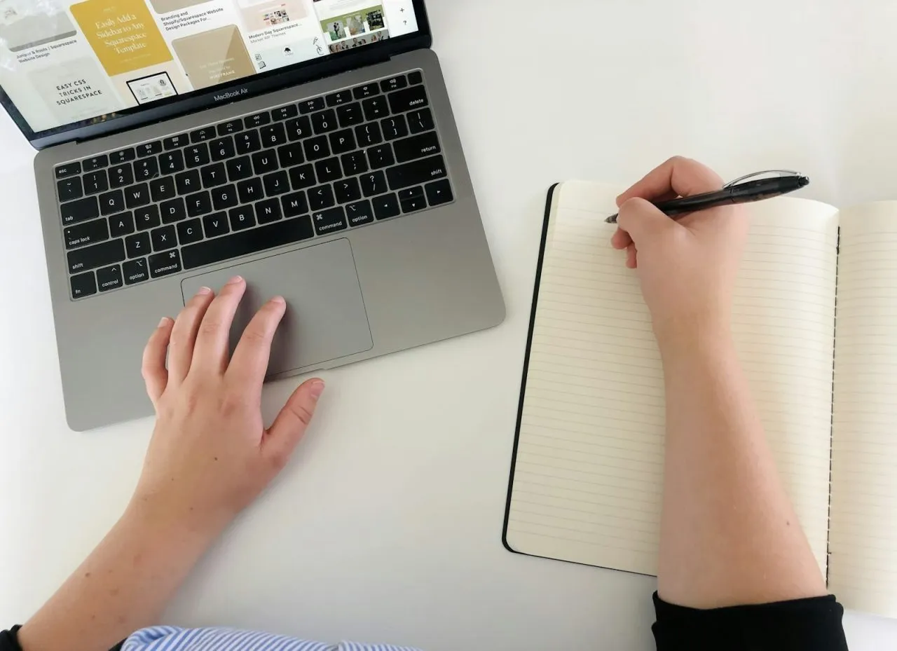 Top view of hands drafting a letter in a notebook next to a laptop