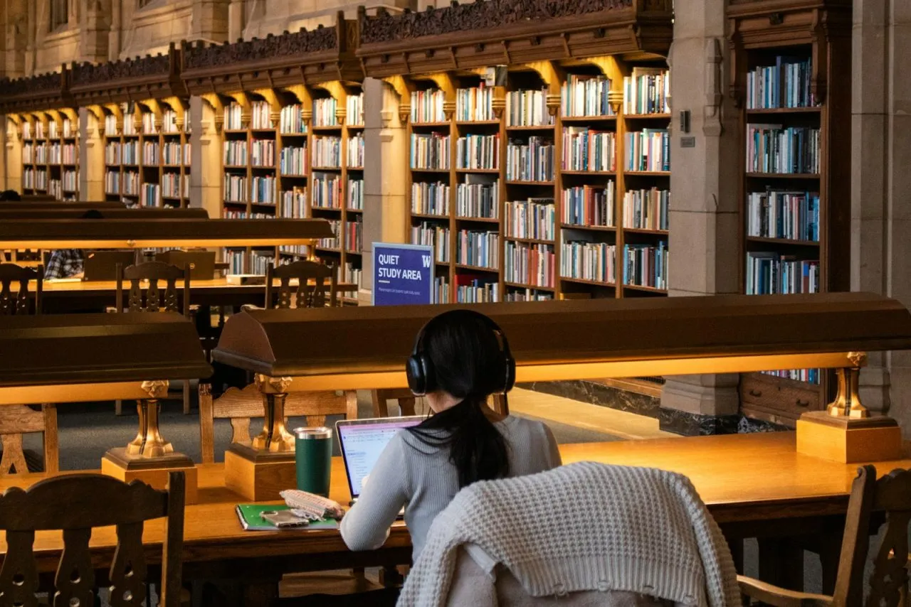 Student wearing headphones studying on a laptop at a long table in a grand library